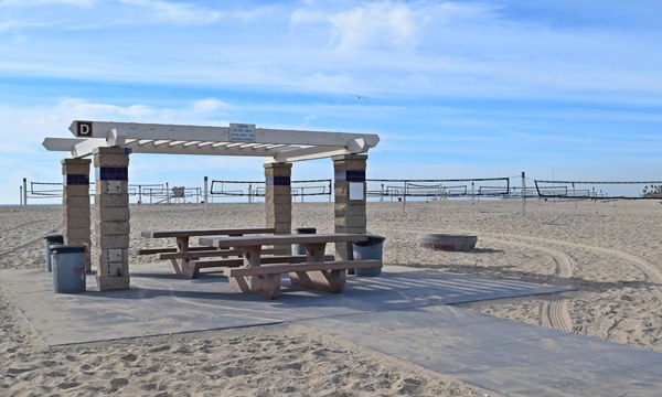Newland Ramada B sits on a paved path (wheelchair accessible). There are two picnic tables and a fire ring adjacent to the area. Volleyball courts line the sand several yards behind the picnic area.